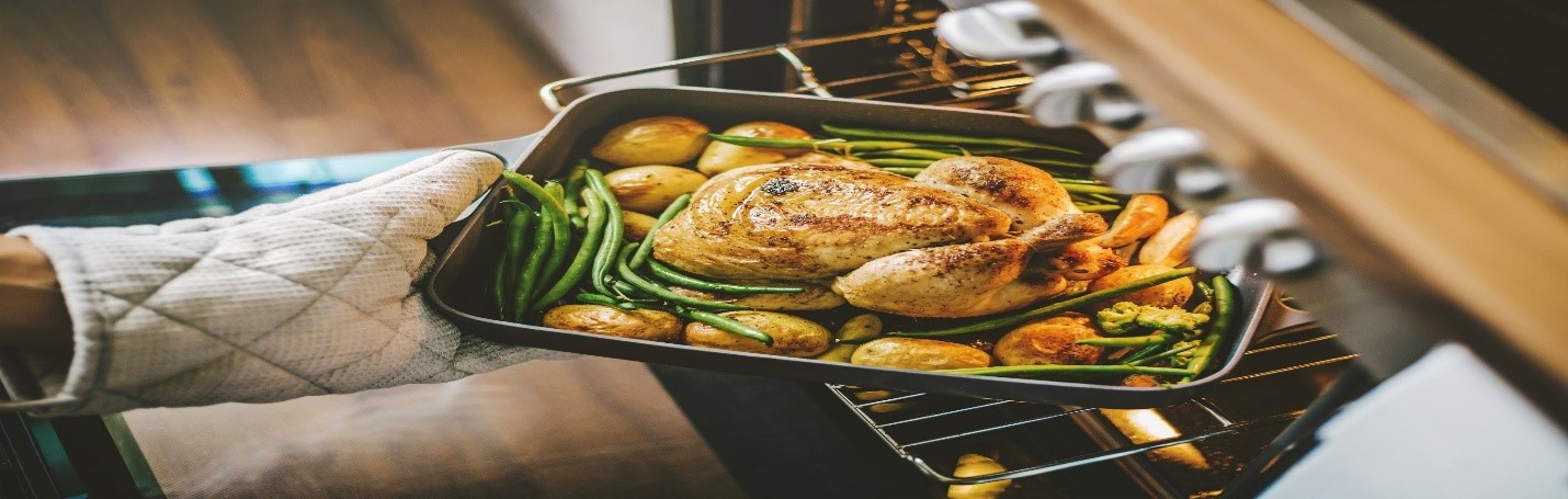 Person wearing oven gloves holding baking tray with cooked whole vegetables and meat over metal rack in open oven