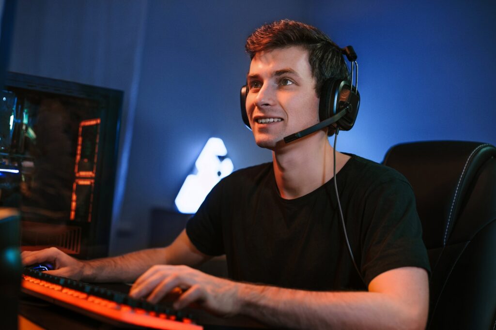 Man wearing black t-shirt and headset sitting in front of orange and black keyboard in dimly lit room with blue walls