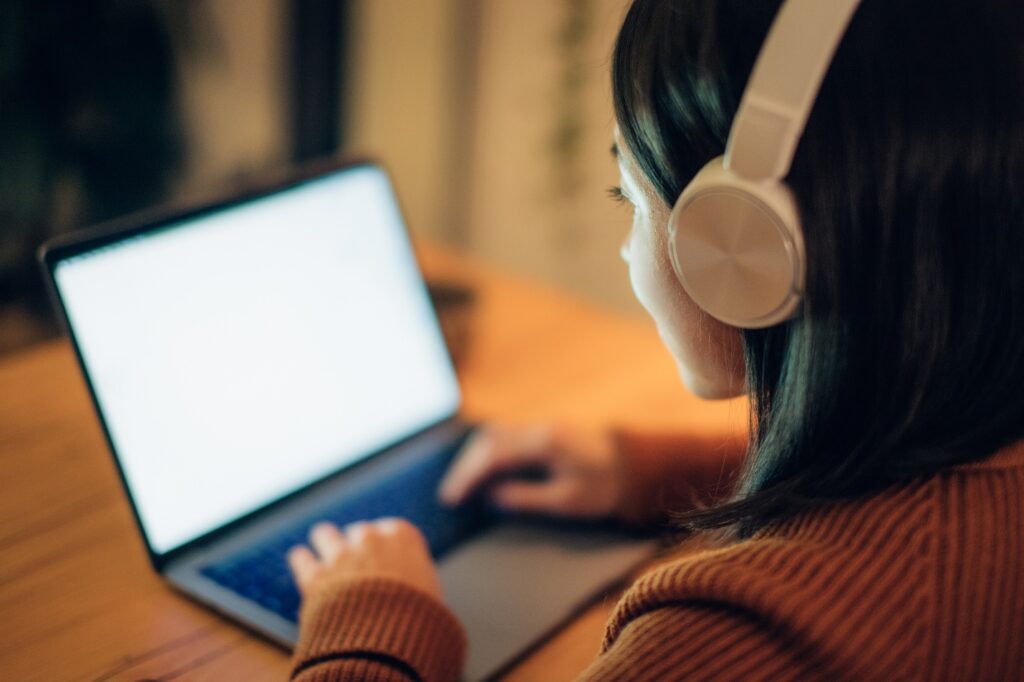 Woman seated at wood table, wearing headphones, working on laptop