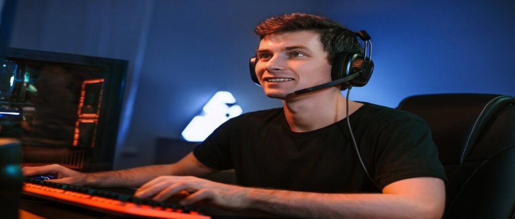 Man wearing black t-shirt and headset sitting in front of orange and black keyboard in dimly lit room with blue walls
