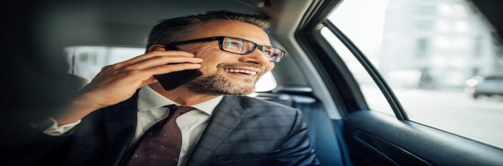 Man with glasses and goatee in business suit in car on back seat, holding cellphone to ear, looking out window and smiling