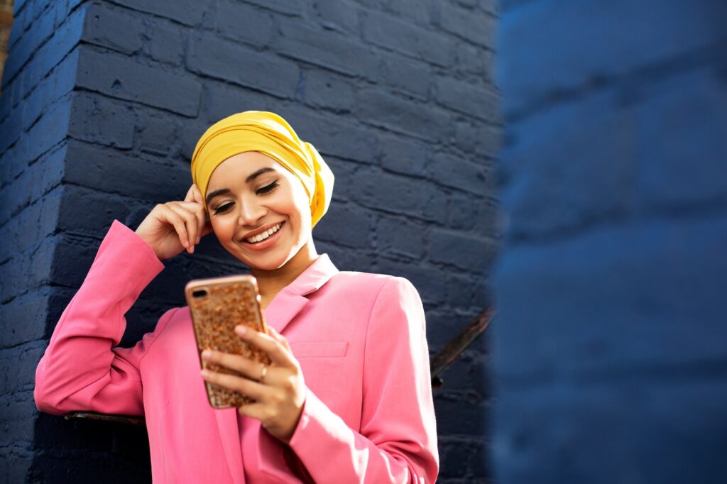 Woman wearing headwrap and jacket, leaning on stair railing at edge of painted brick wall, holding and looking at cellphone