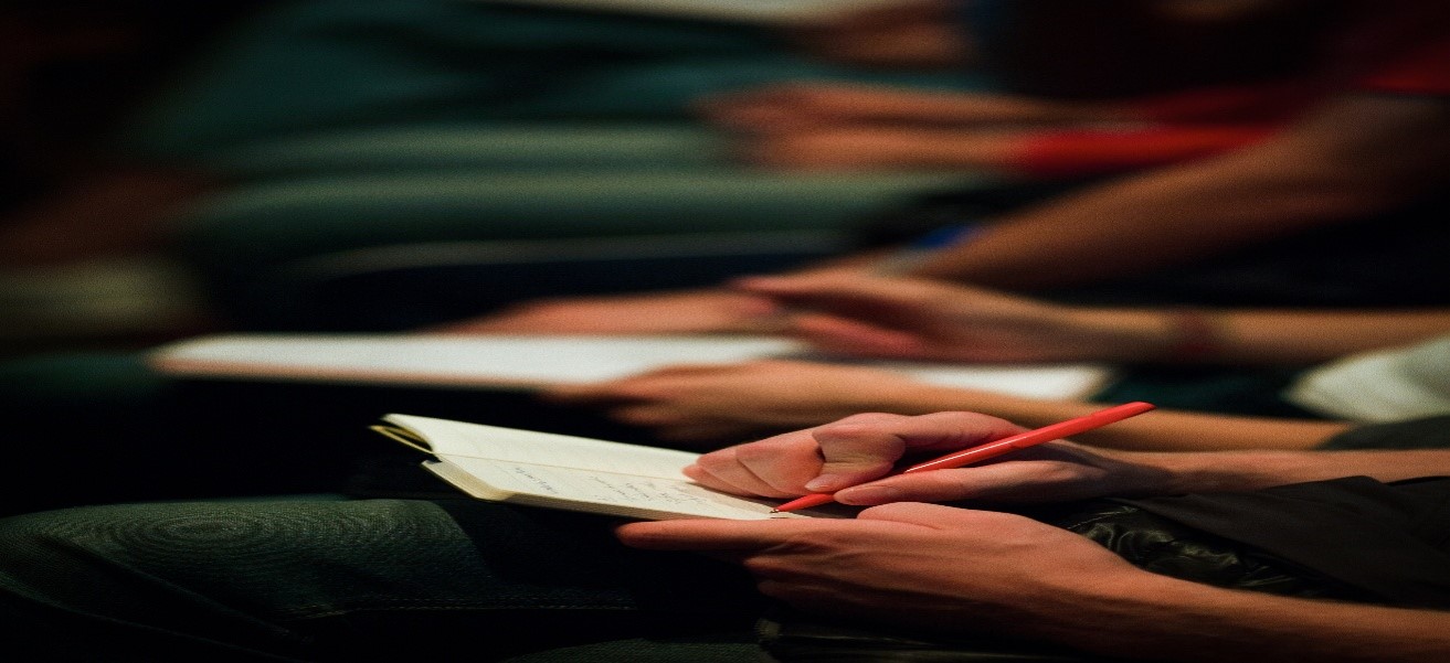 Row of seated people, some writing notes in book on lap