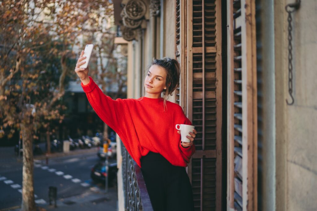 Smiling woman standing on balcony, with mug in hand, holding cellphone to take photo of self