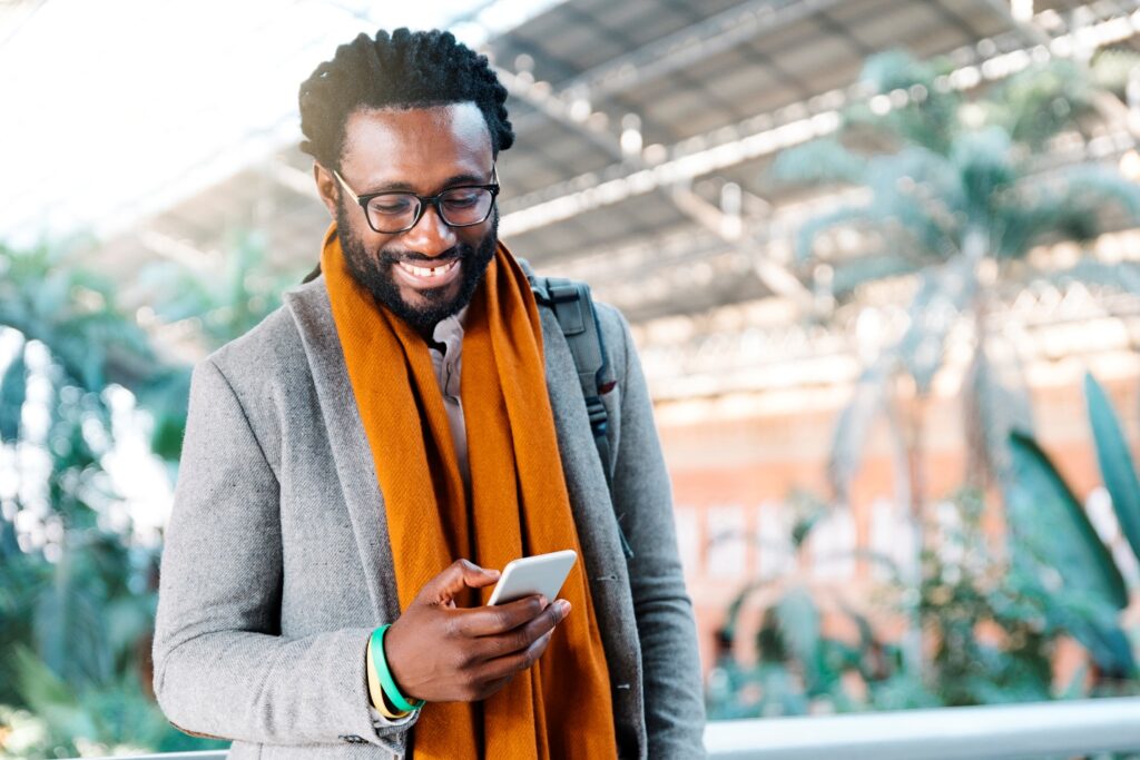 Smiling man wearing coat and scarf, with bag strap on shoulder, holding and looking at cellphone